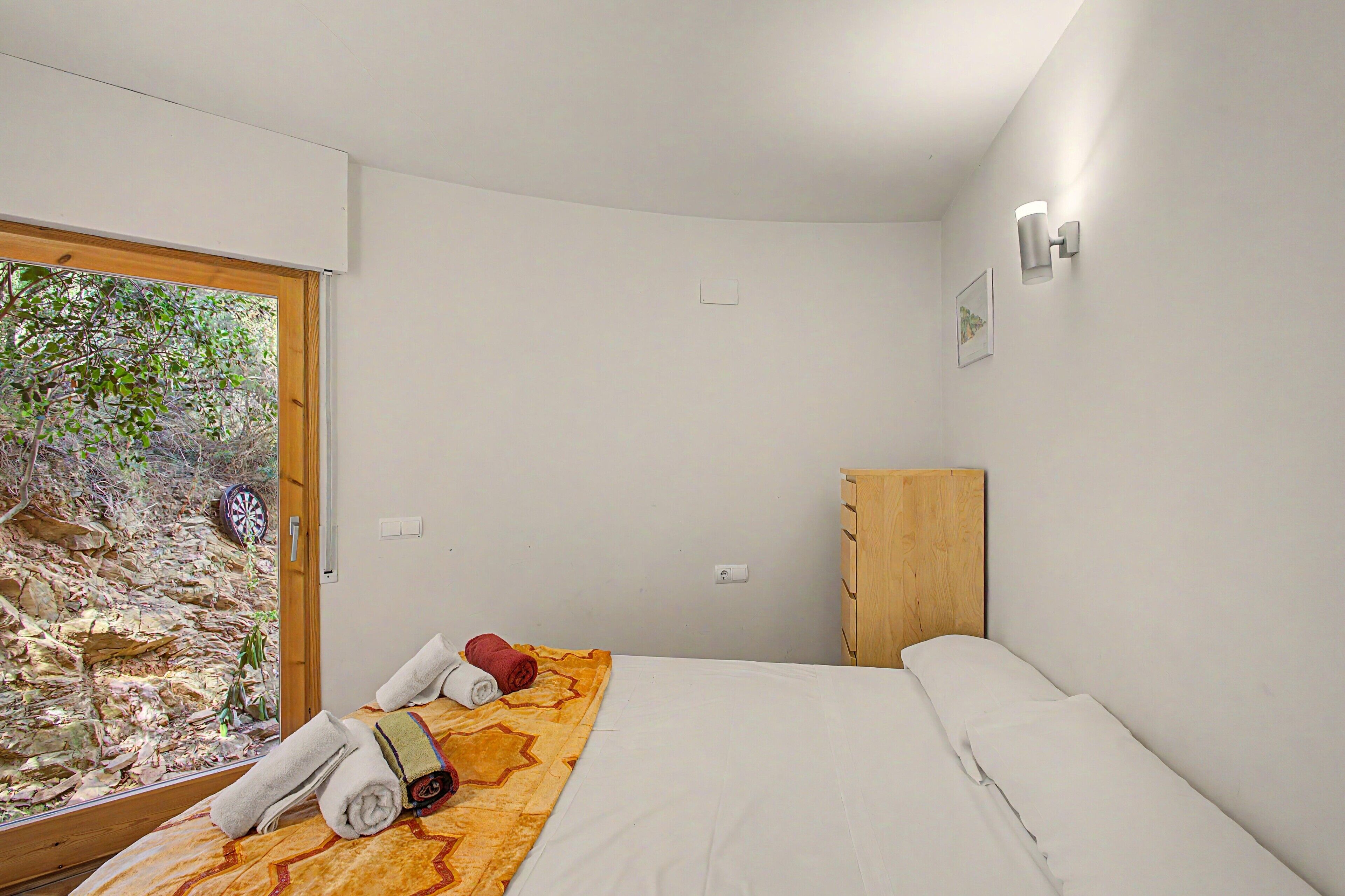 A bedroom with a bed, a wooden wardrobe, a window with a view of trees, and a door.