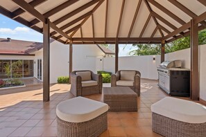 Überdachte Terrasse im Freien mit Korbmöbeln, einem Grill und Blick auf ein Haus im Hintergrund.