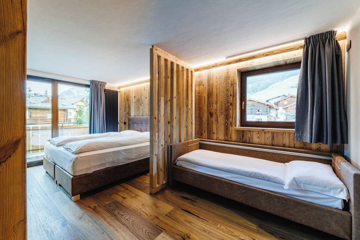 A room with two wooden beds, a large window with curtains, and a view of buildings outside.
