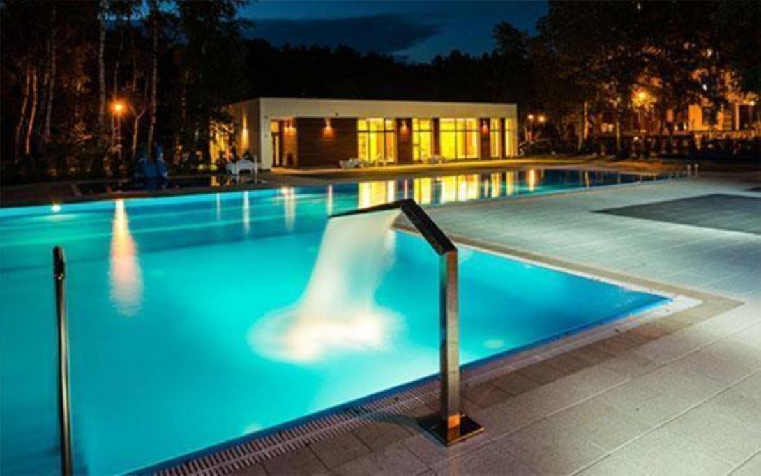 Ein Schwimmbecken mit klarem Wasserspiel, umgeben von einer gepflasterten Liegefläche und einem Gebäude mit großen Fenstern.