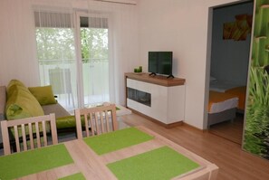 Une pièce comprenant un canapé, une table à manger avec des sets de table verts, un téléviseur posé sur un meuble et un lit avec une tête de lit blanche.