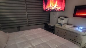 A bedroom with a bed, a desk with a printer, a television displaying a fire, and a digital clock. - The River Condos (Detroit)