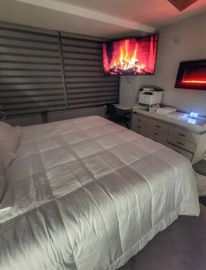 A bedroom with a bed, a desk with a printer, a television displaying a fire, and a digital clock. - The River Condos (Detroit)