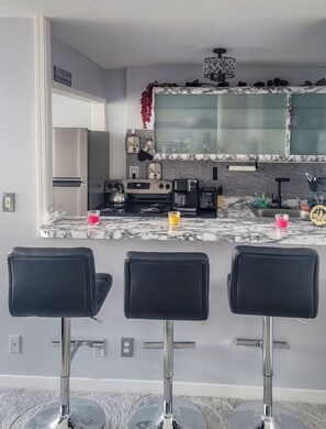 Una cocina moderna con un refrigerador de acero inoxidable, una cafetera y un hervidor en la encimera. Hay tres taburetes negros frente a la encimera.