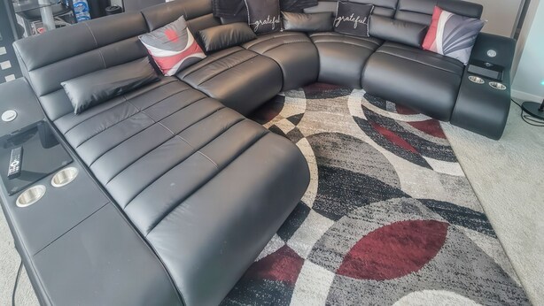 A modern living room with a sectional sofa, a chandelier, and a kitchen in the background. - The River Condos (Detroit)