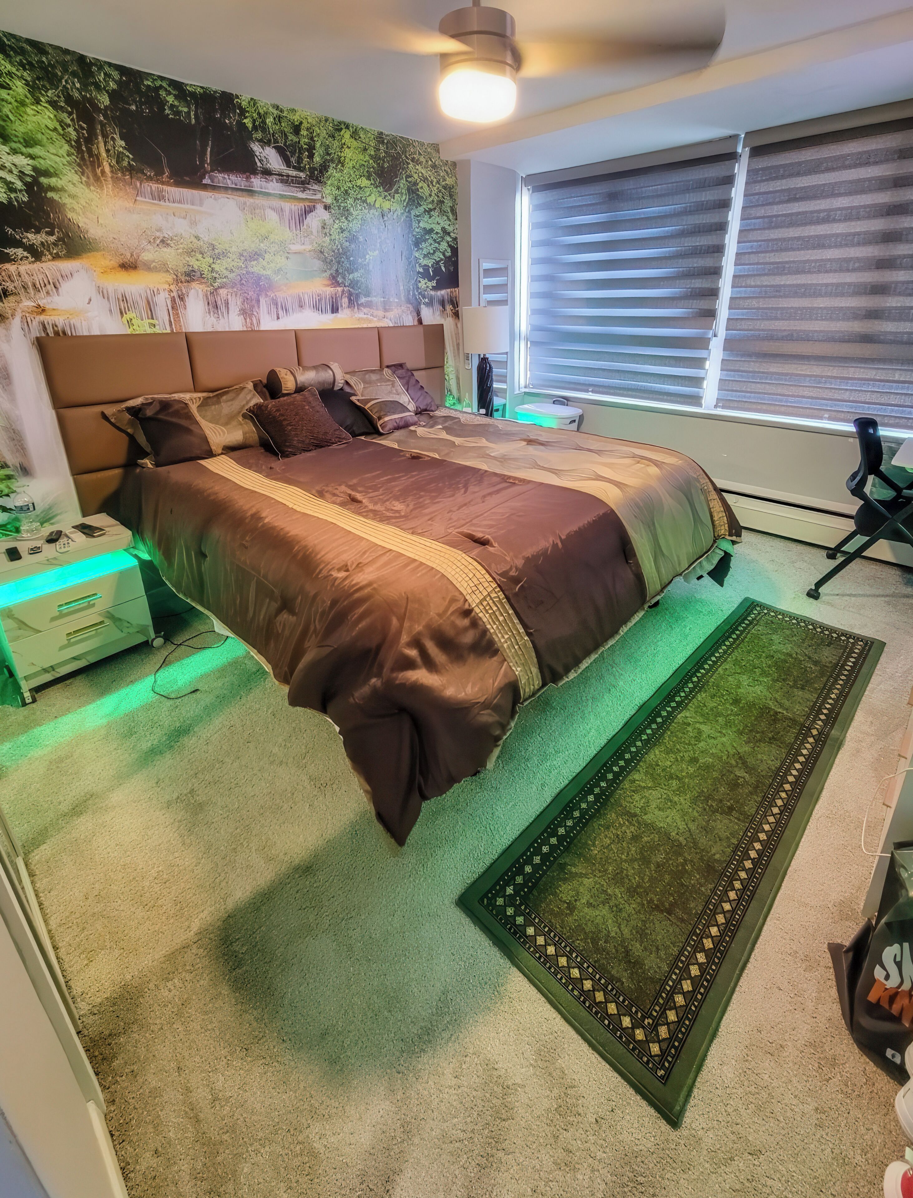 A bedroom with a bed, a nightstand, a lamp, a window with blinds, and a wall mural.