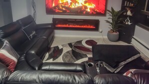A living room with a leather sofa, a flat-screen TV, a fireplace, and a chandelier. - The River Condos (Detroit)