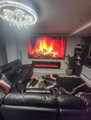 A living room with a leather sofa, a flat-screen TV, a fireplace, and a chandelier. - The River Condos (Detroit)