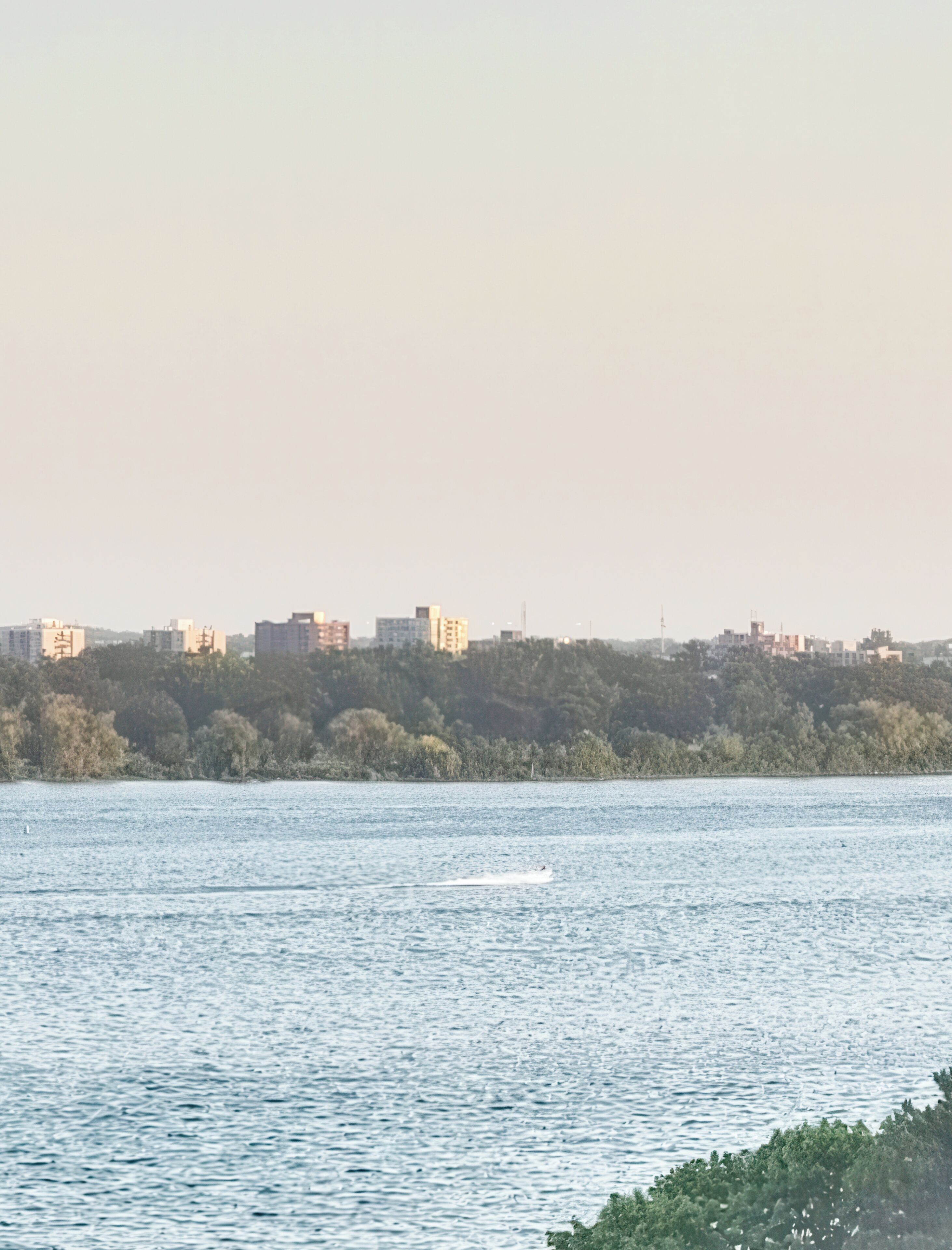 Ein Gewässer mit einem sich bewegenden Boot. Im Hintergrund eine Stadtansicht mit Gebäuden und einem bewaldeten Hügel.