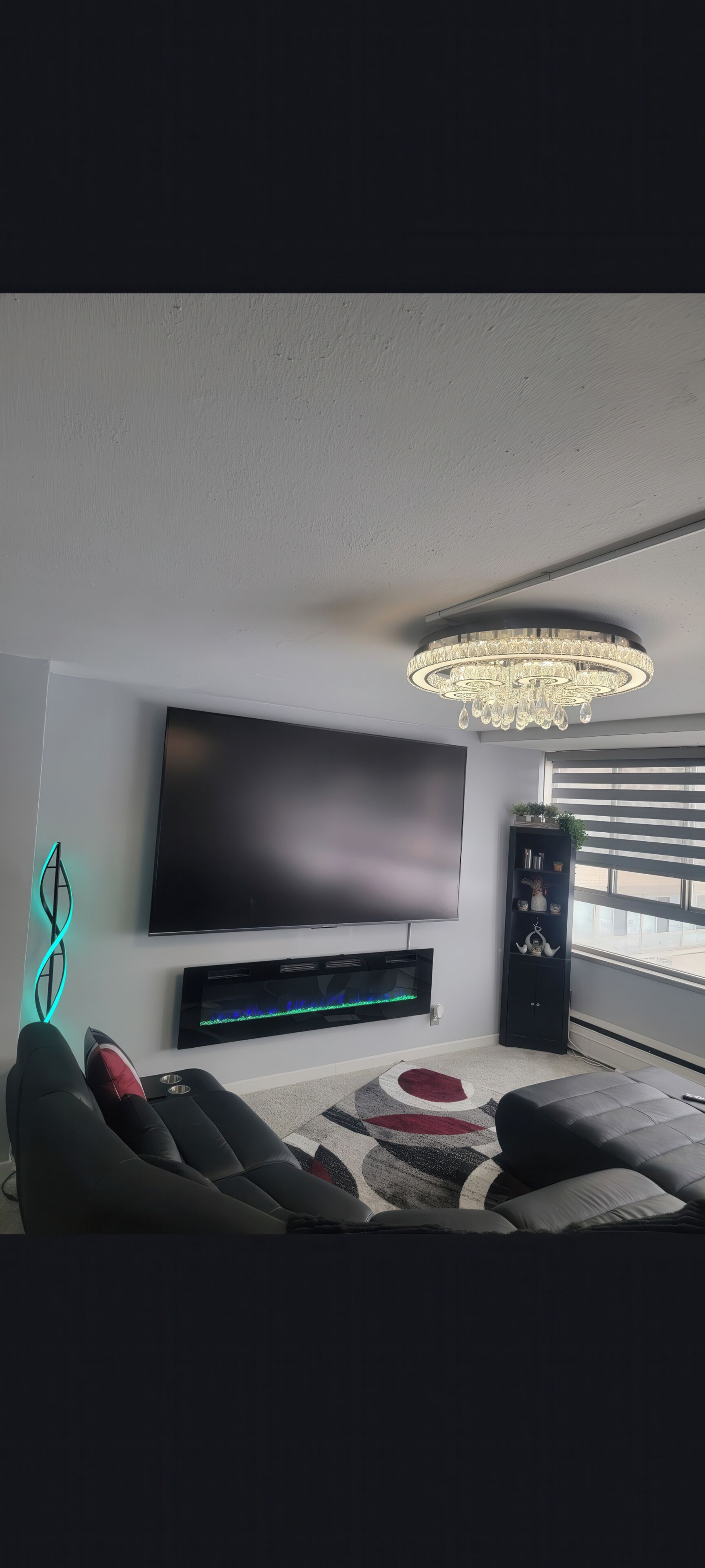 A modern living room with a flat-screen TV, a soundbar, a decorative chandelier, a black entertainment unit, and a patterned rug.