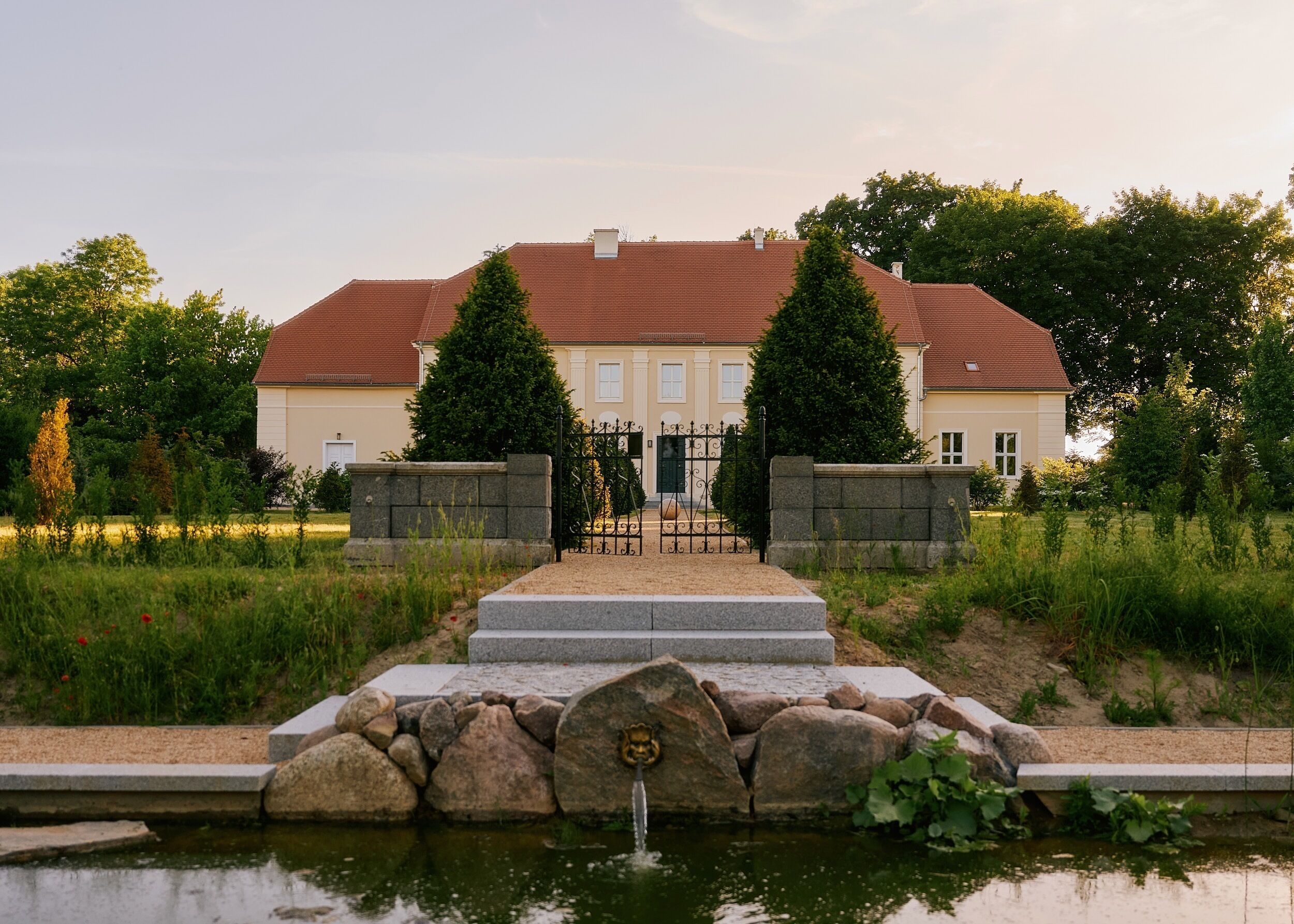 Ein großes, zweistöckiges Gebäude mit rotem Ziegeldach, einer Steinmauer und einem Teich mit einer Fontäne im Vordergrund.