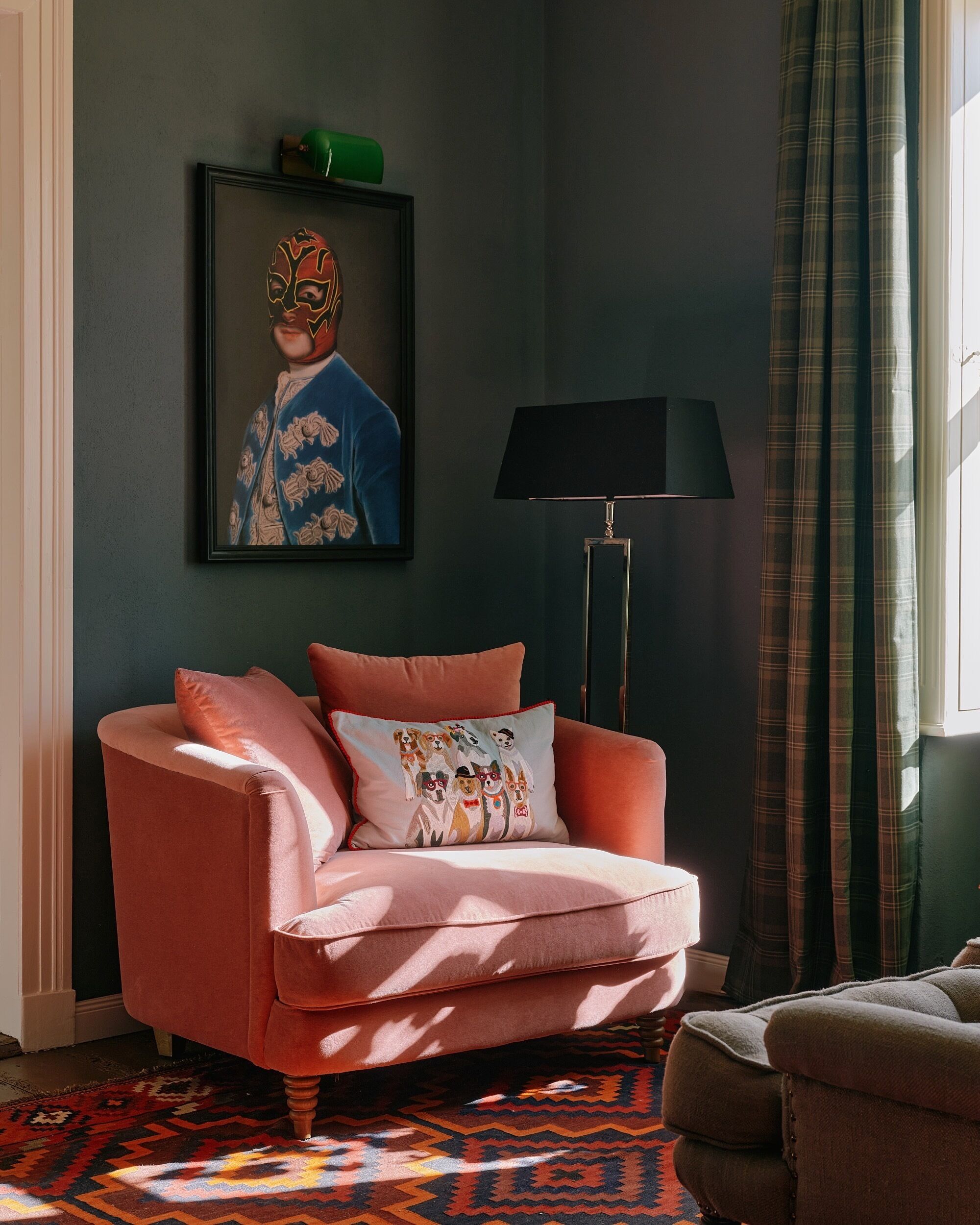 Een kamer met een roze fauteuil, een lijst met een portret, een vloerlamp en een geruit gordijn.