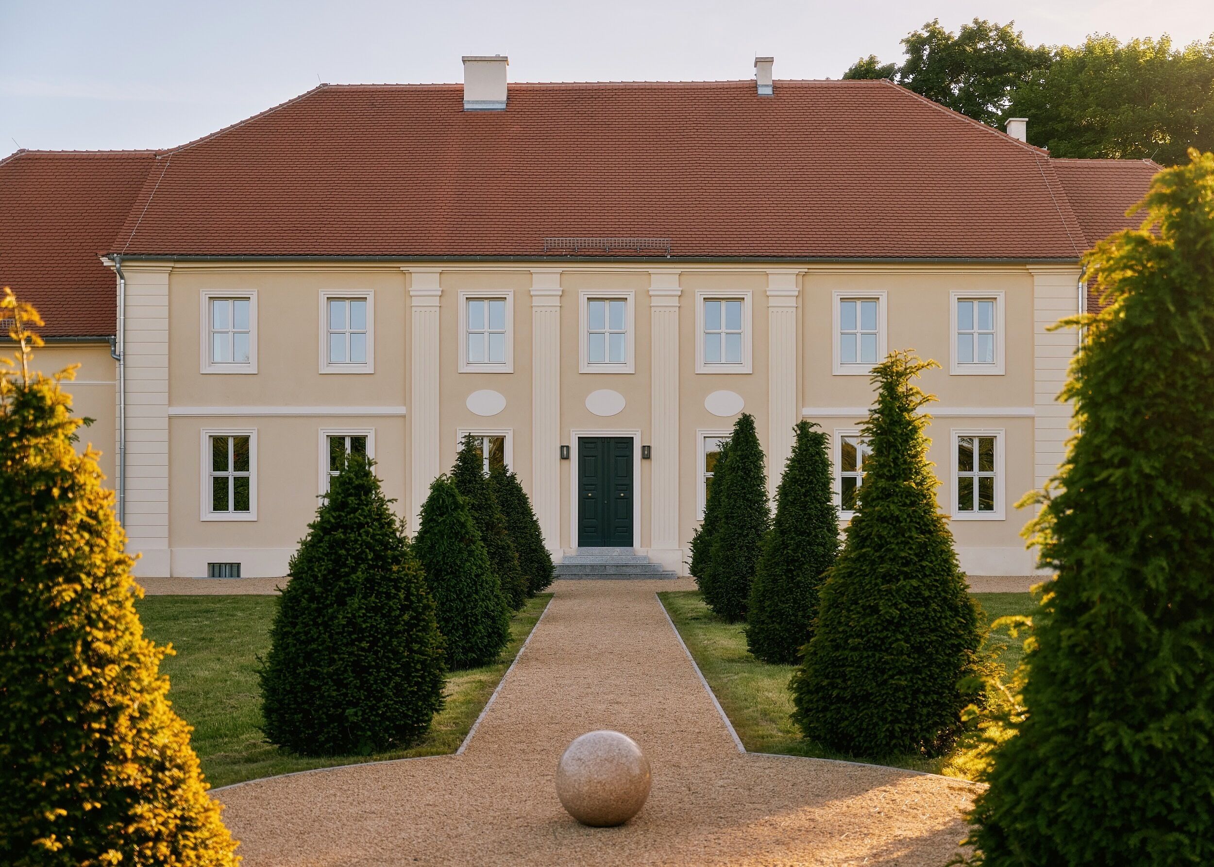 Ein großzügiges, symmetrisches Gebäude mit einer zentralen Eingangspforte, umgeben von einem Kiesweg und formal geschnittenen Hecken.