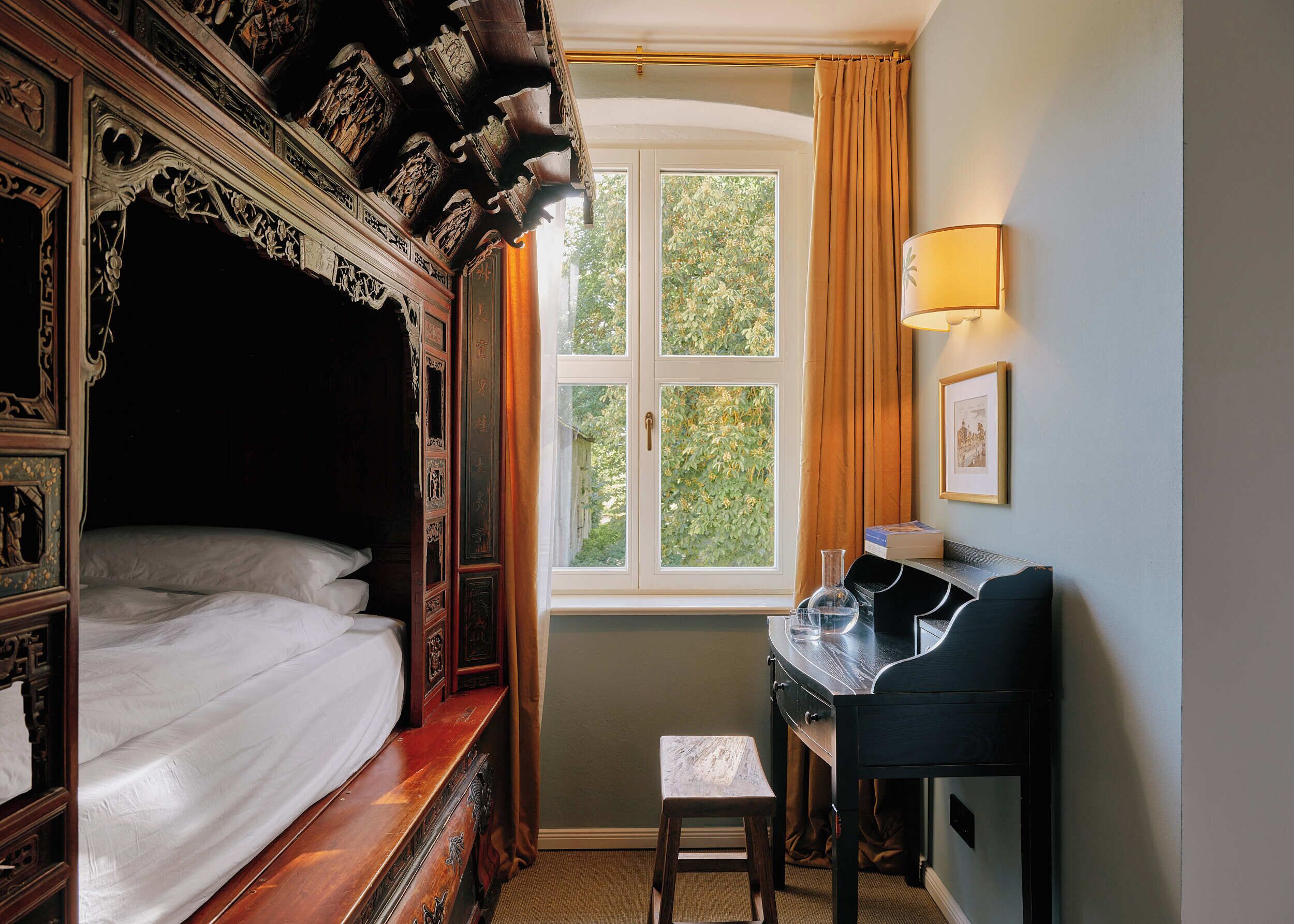 Een kamer met een hemelbed, een bureau met stoel, een klein tafeltje en een raam met gordijnen.