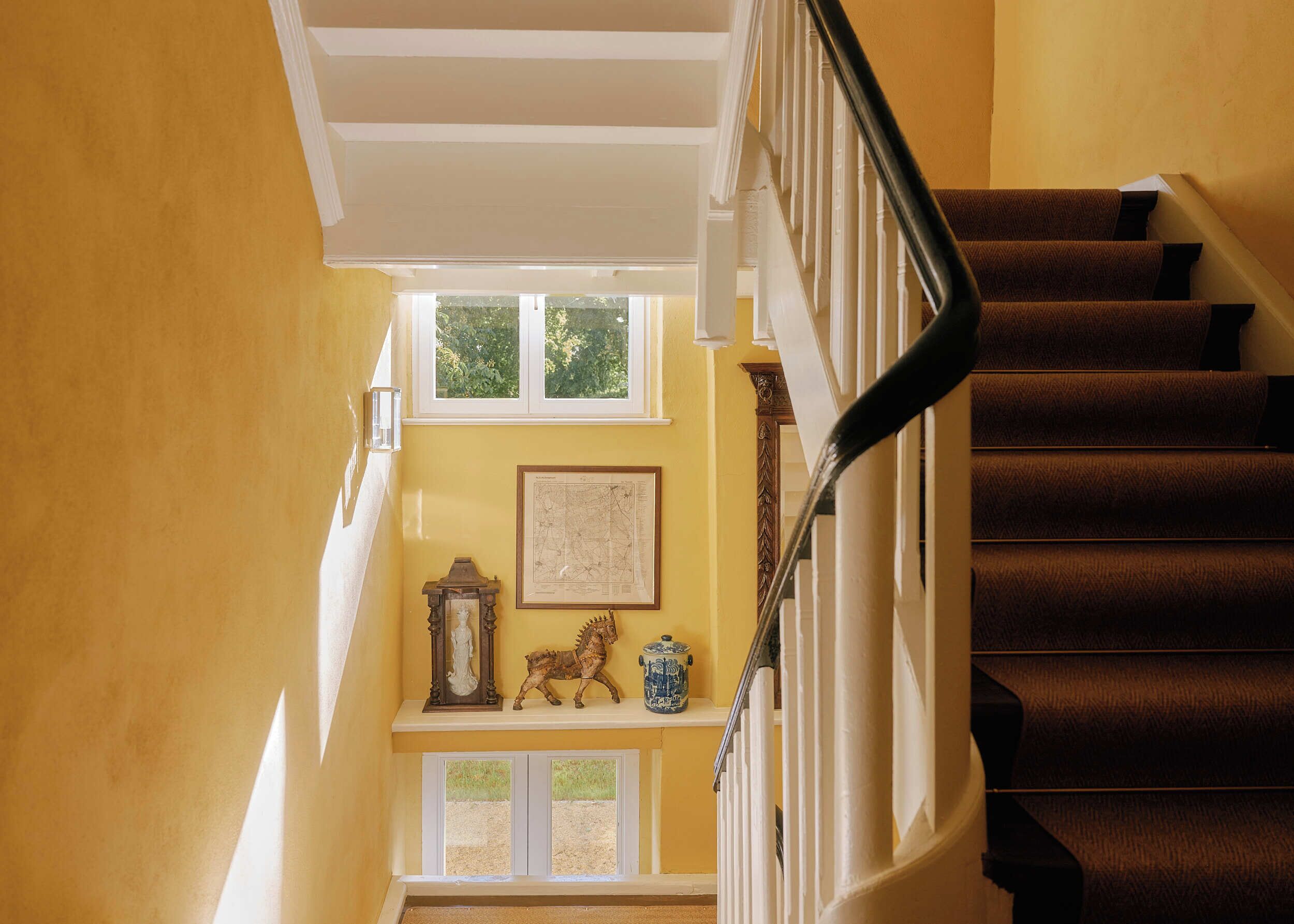 A staircase with a dark handrail, a white balustrade, and a yellow wall with a framed map and decorative items.