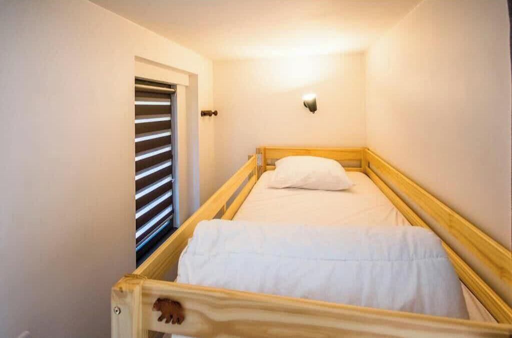 Une petite chambre avec un lit simple, un cadre en bois, un matelas blanc et un luminaire fixé au mur.