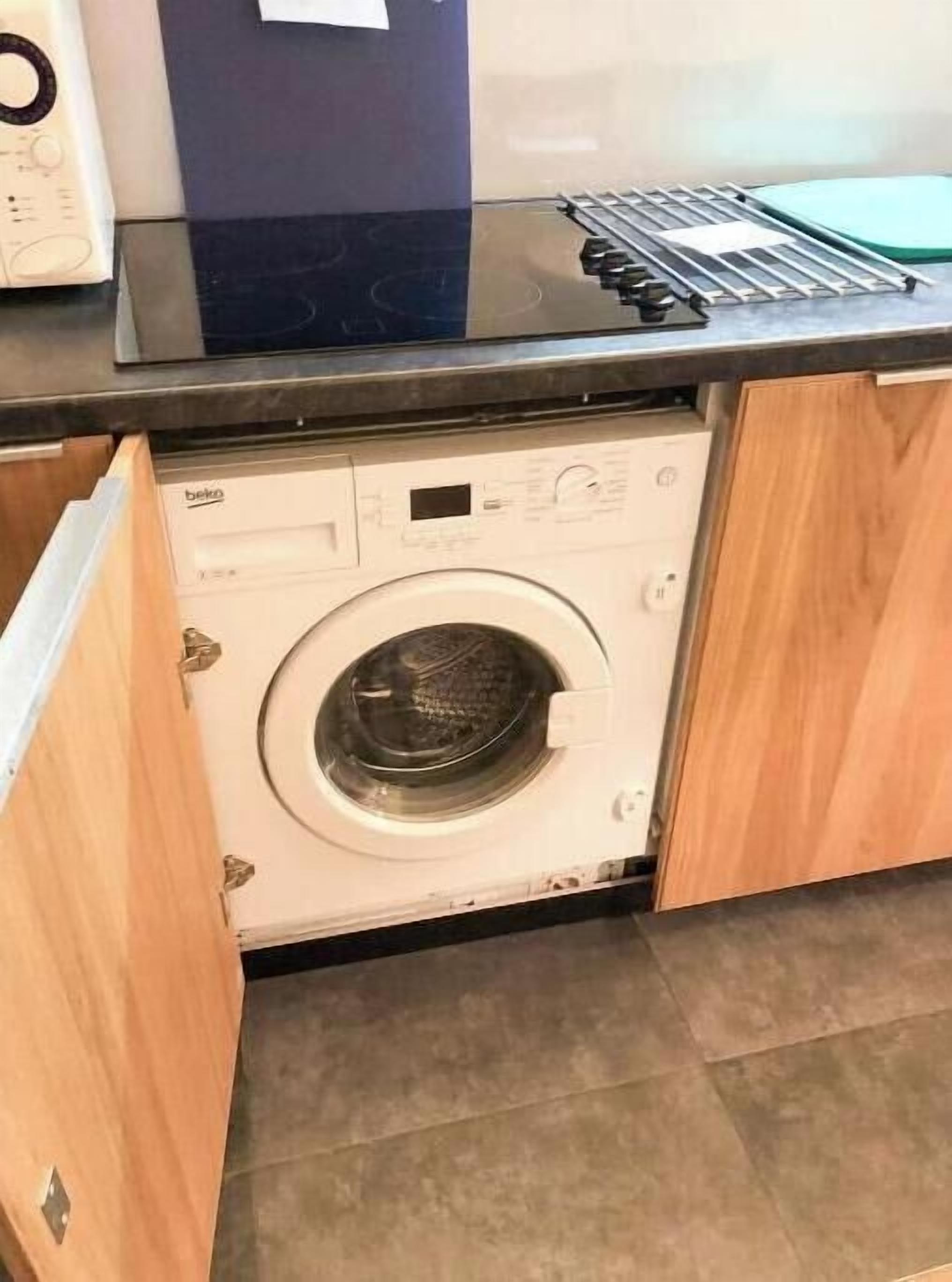 Un lave-linge compact dans une petite cuisine, avec des meubles en bois et un sol carrelé.