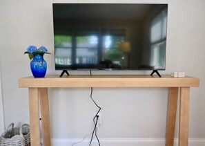 A television on a wooden stand with a vase of flowers and a stack of books. - Raynor Shine | Walk to Beach | Family Fun | Dog Friendly (Falmouth)