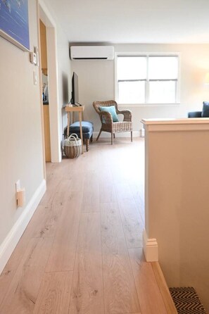 A hallway with a wooden floor, a wicker chair, a television on a stand, and a window with blinds. - Raynor Shine | Walk to Beach | Family Fun | Dog Friendly (Falmouth)