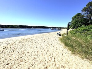 A sandy beach with a clear blue sky, a few boats on the water, and a chair on the sand. - Raynor Shine | Walk to Beach | Family Fun | Dog Friendly (Falmouth)