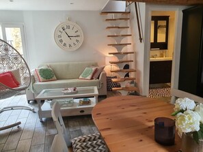 A living room with a sofa, coffee table, and a wooden dining table. There is a wall clock and a staircase leading to another room. - Traverse du Vieux Château (Menton)