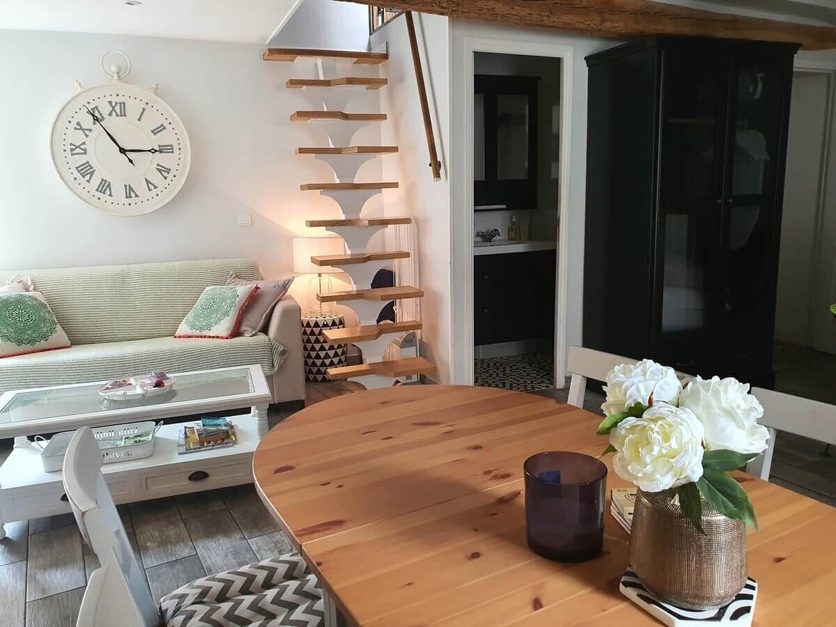 A cozy living room with a wooden dining table, a sofa with cushions, a wall clock, and a staircase.