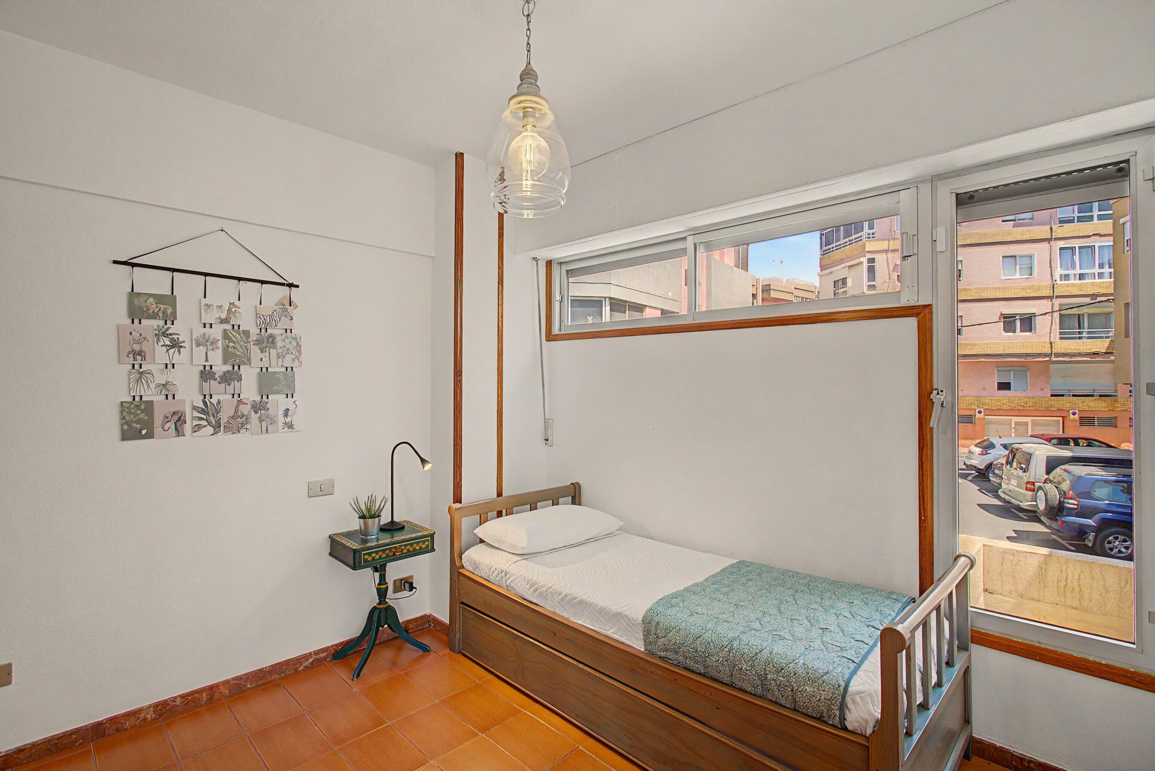 Una habitación pequeña con cama individual, estructura de madera, lámpara colgante, planta en la pared y ventana con vista a un estacionamiento y edificios.