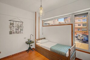 Een kleine eenpersoonskamer met een houten bedframe, een hanglamp, een aan de muur bevestigde plant en een raam met uitzicht op een parkeerplaats en gebouwen.