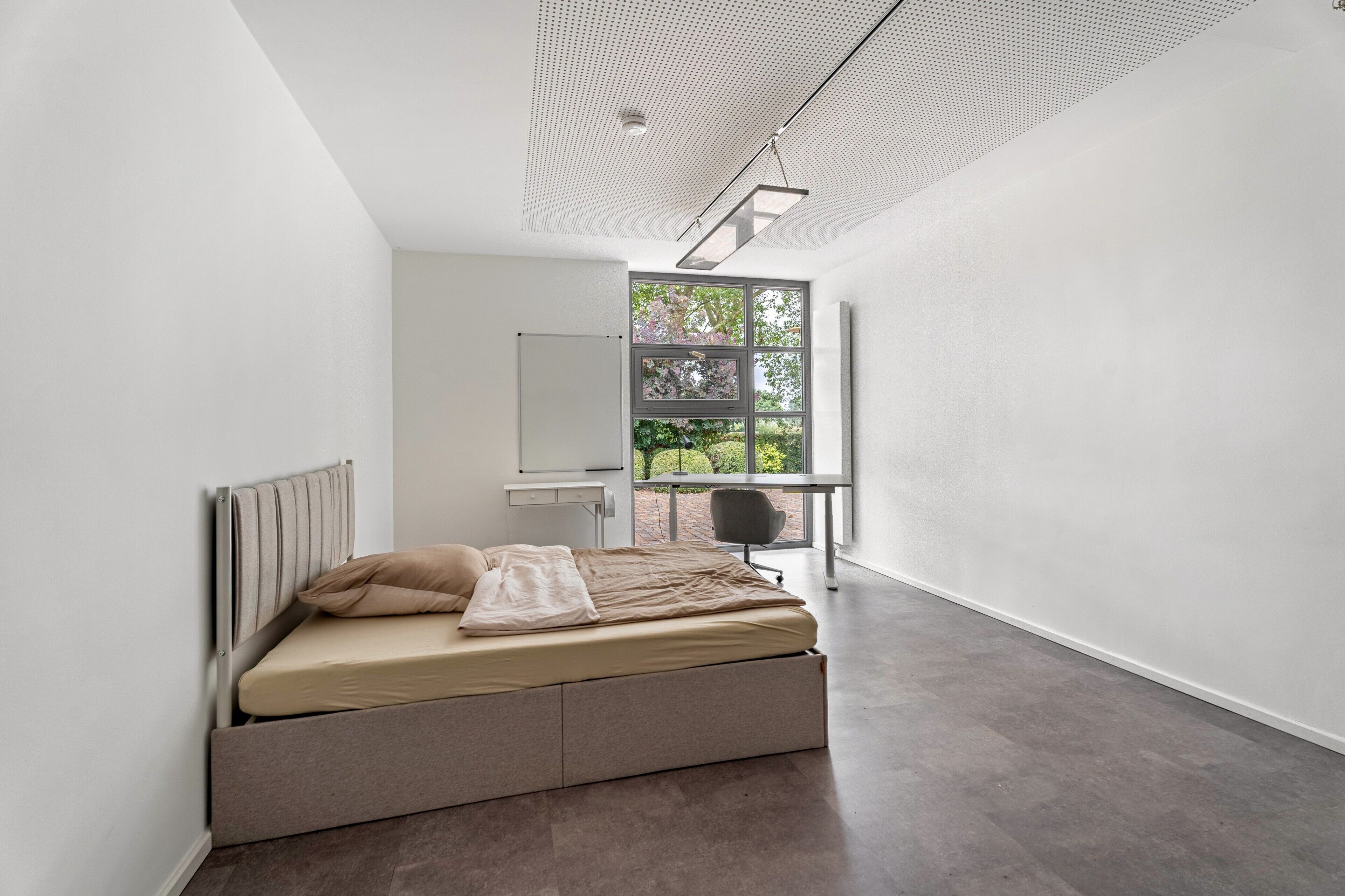 A minimalist bedroom with a bed, a desk, and a view of greenery through a window.