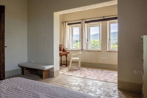 Un dormitori amb un llit, una banca, un escriptori i dues finestres amb cortines.