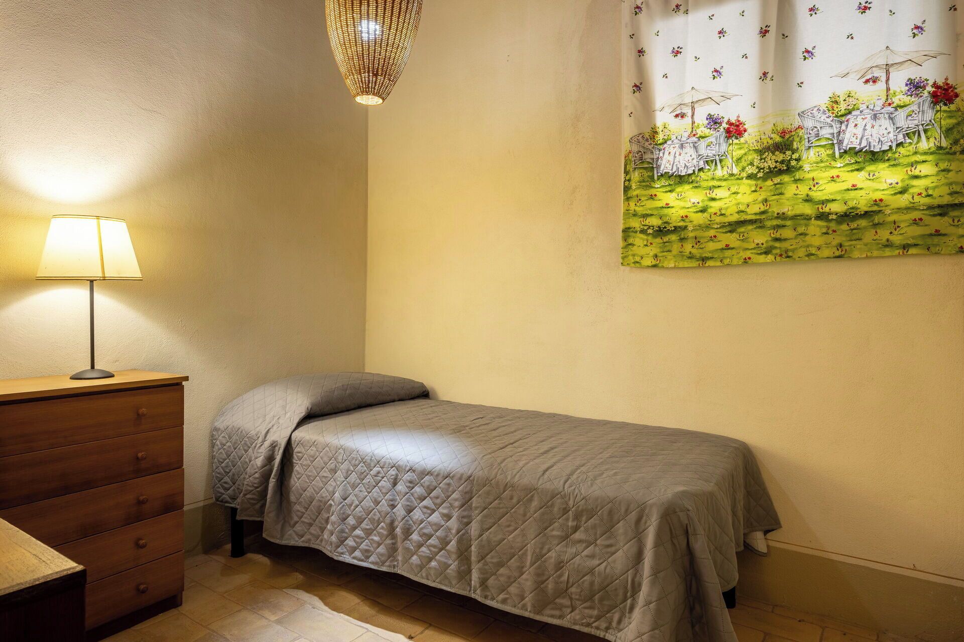 A single bed with a quilted cover, a wooden dresser with drawers, a table lamp, and a wall-mounted painting of a garden scene.