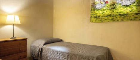 A single bed with a quilted cover, a wooden dresser with drawers, a table lamp, and a wall-mounted painting of a garden scene.