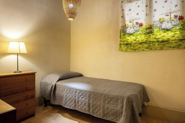 A single bed with a quilted cover, a wooden dresser with drawers, a table lamp, and a wall-mounted painting of a garden scene.