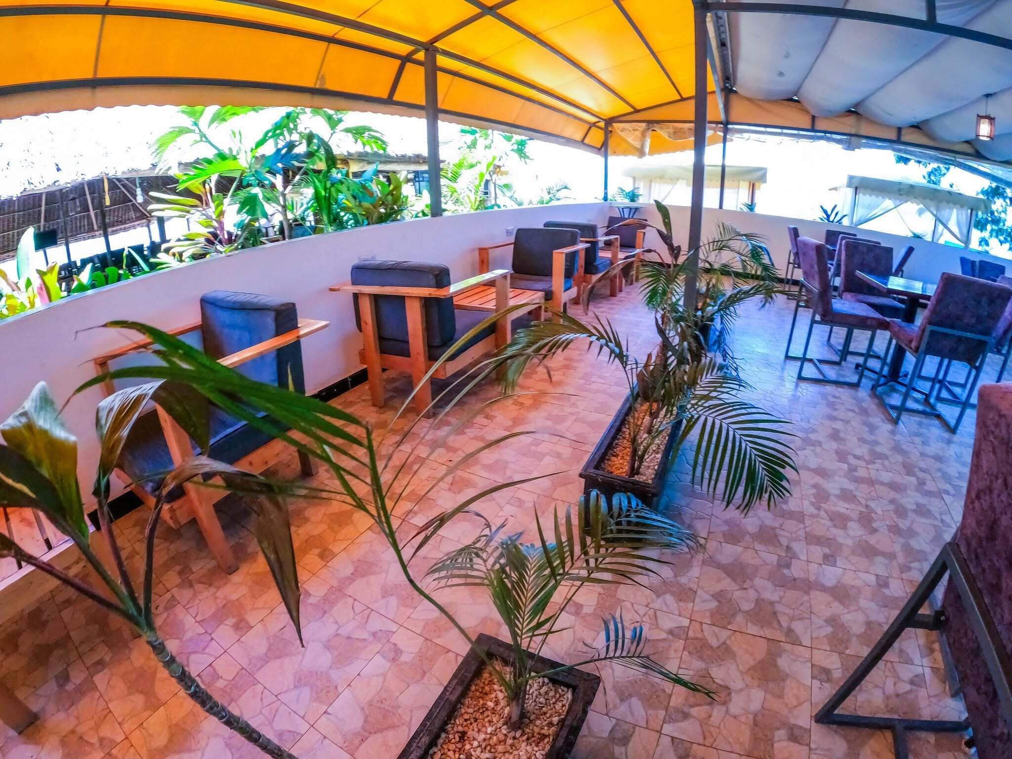 An outdoor seating area with tables and chairs, surrounded by potted plants and a yellow canopy.