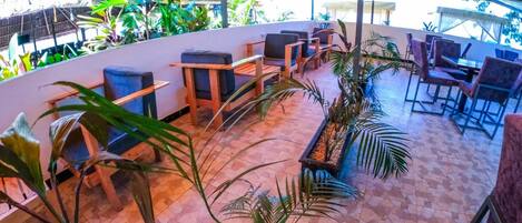 Un espace extérieur aménagé avec des tables et des chaises, entouré de plantes en pot et surmonté d’un auvent jaune.