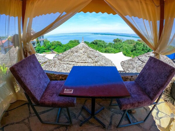 A table with two chairs under a canopy with a view of a beach and thatched-roof buildings.