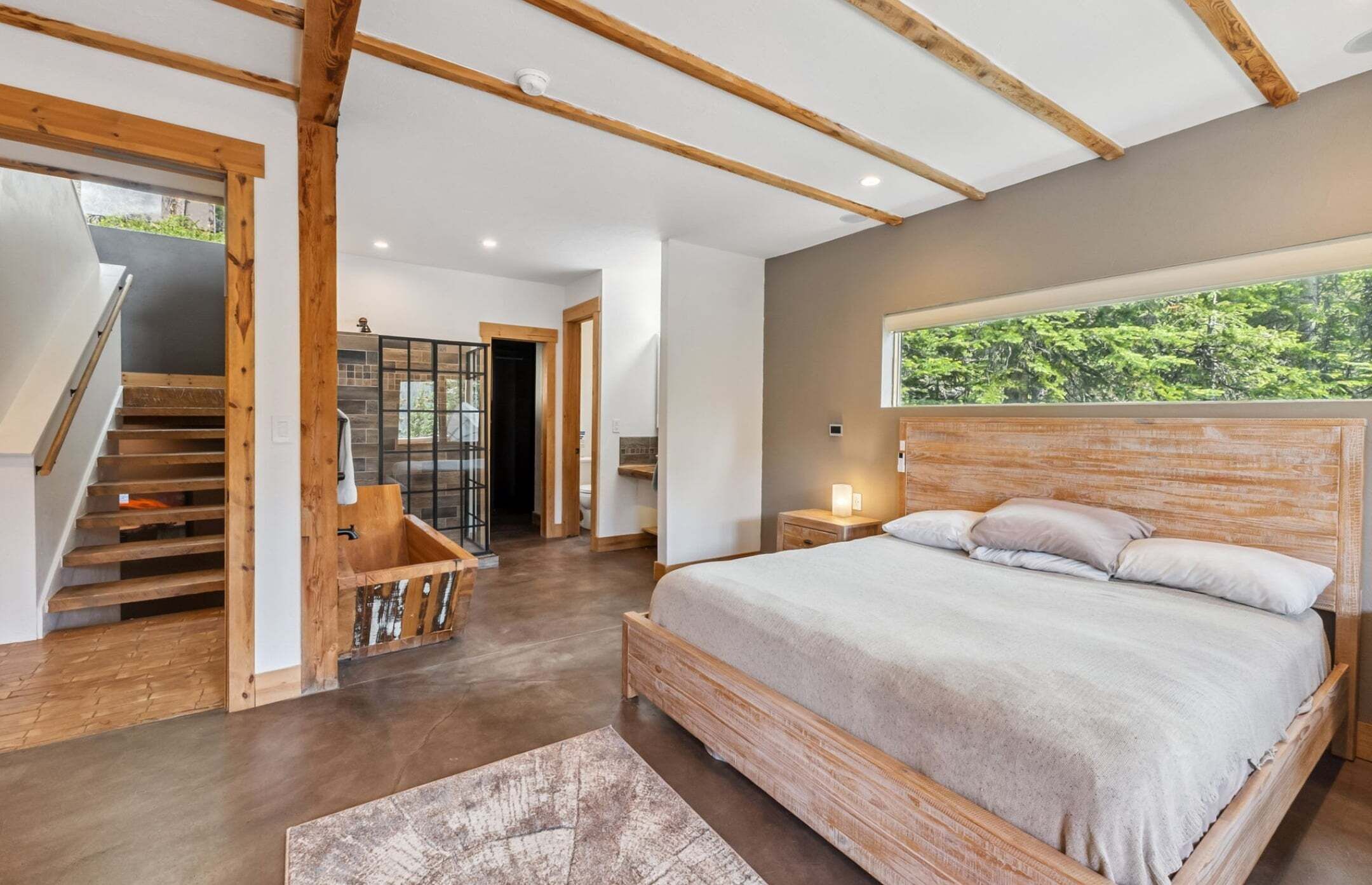A bedroom with a large bed, wooden beams, a view of greenery, and a staircase.