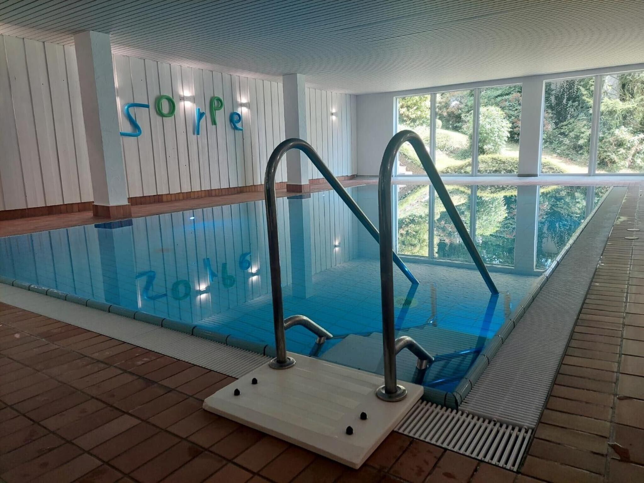 Ein Hallenbad mit großen Glaswänden, einer Holzterrasse und Blick auf Grünflächen.