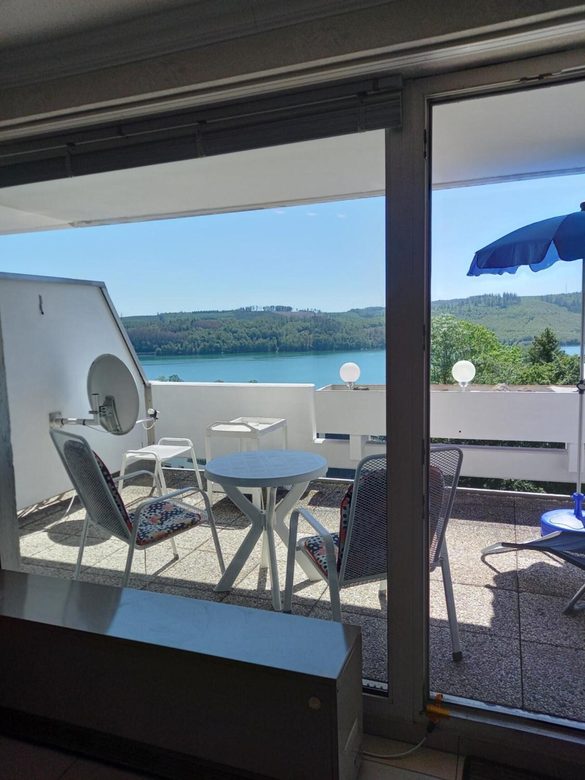 Ein Balkon mit Tisch und Stühlen, einem blauen Sonnenschirm und Blick auf einen See und Hügel.