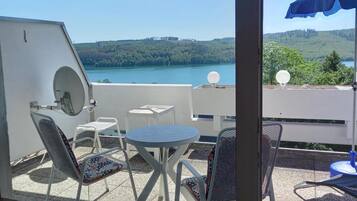 Ein Balkon mit Tisch und Stühlen, einem blauen Sonnenschirm und Blick auf einen See und Hügel.