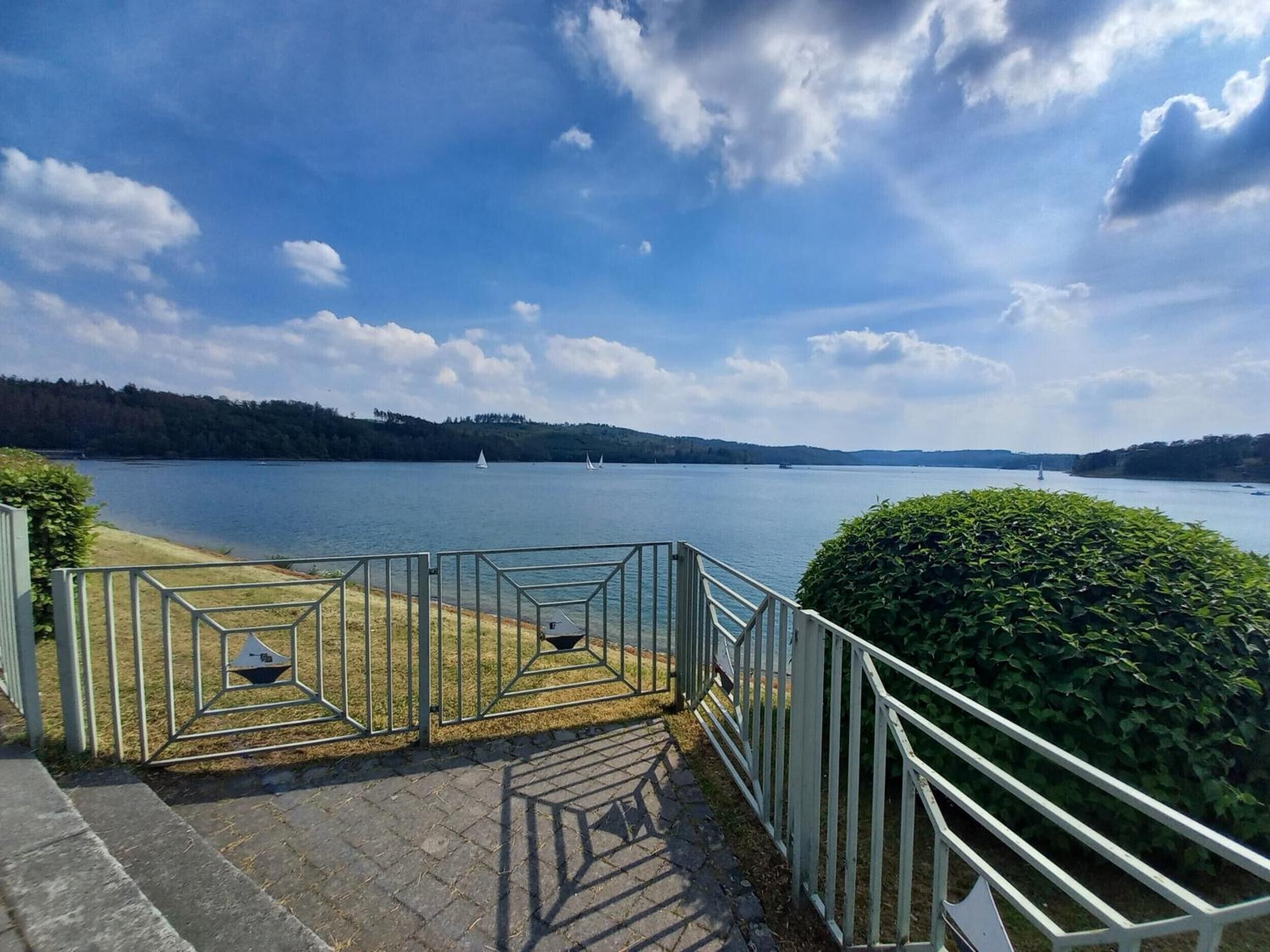 Ein gepflasterter Weg mit Metallgeländern führt zu einem See mit Segelbooten.