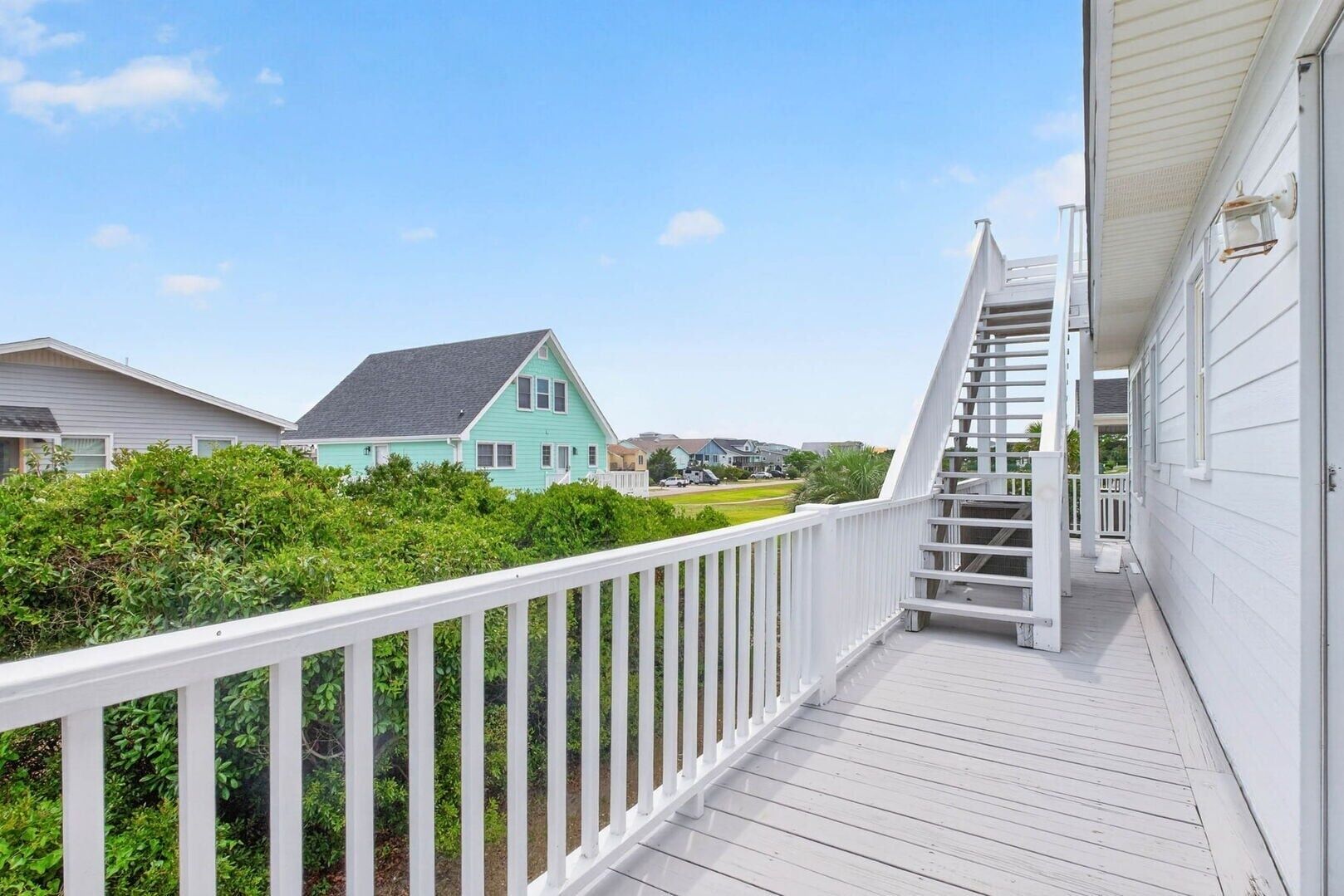 A wooden deck with a white railing, a staircase leading to a porch, and a turquoise house in the background.