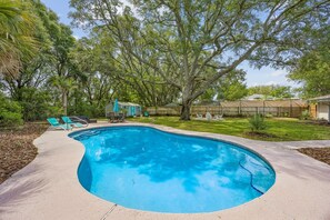 Ein Pool mit gewölbter Kante, umgeben von einer Betonplattform. Liegestühle und ein kleiner Tisch mit Sitzgelegenheiten sind vorhanden.
