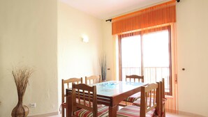 A dining area with a glass-top table, wooden chairs, a vase with dried plants, and a window with orange curtains. - MyStay - Apartment in Alvor with a pool (Alvor)