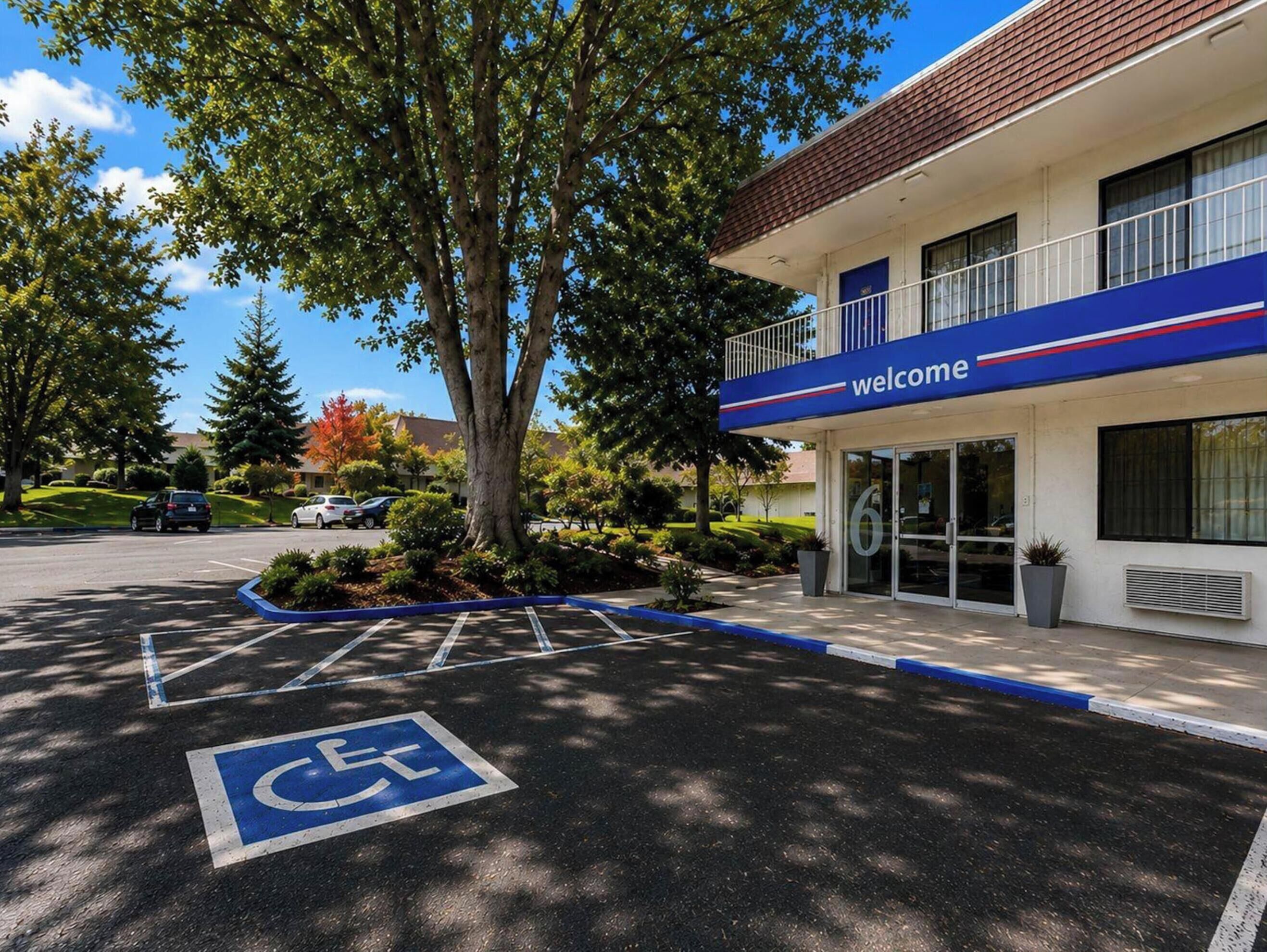 Un bâtiment avec une pancarte indiquant « Bienvenue », un parking avec une place réservée aux personnes handicapées et un arbre devant.