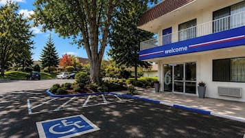 Un bâtiment avec une pancarte indiquant « Bienvenue », un parking avec une place réservée aux personnes handicapées et un arbre devant.