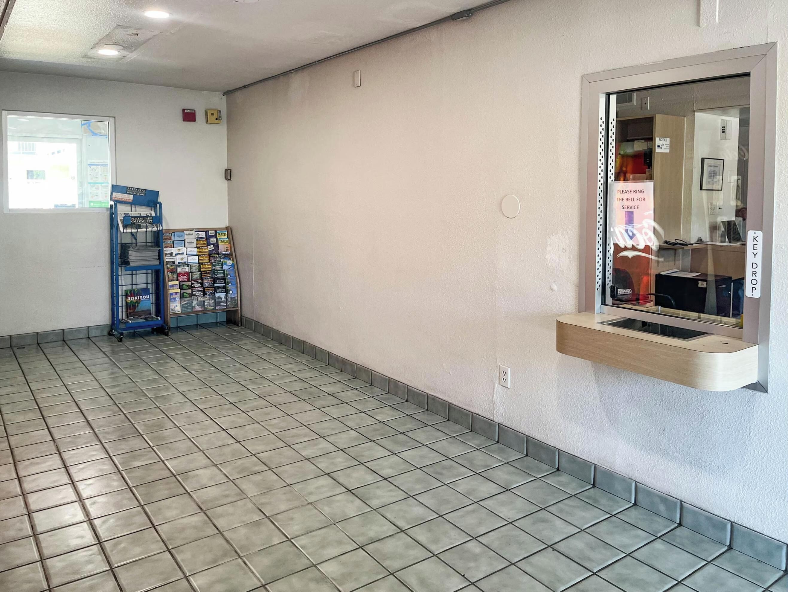 Eine saubere, geflieste Lobby mit einer Rezeption, einem Schwarzesbrett mit verschiedenen Aushängen und einem Fenster mit Blick nach draußen.