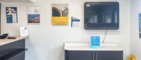 A reception area with a desk, a trash bin, a caution sign, a mounted TV, and informational posters on the wall.