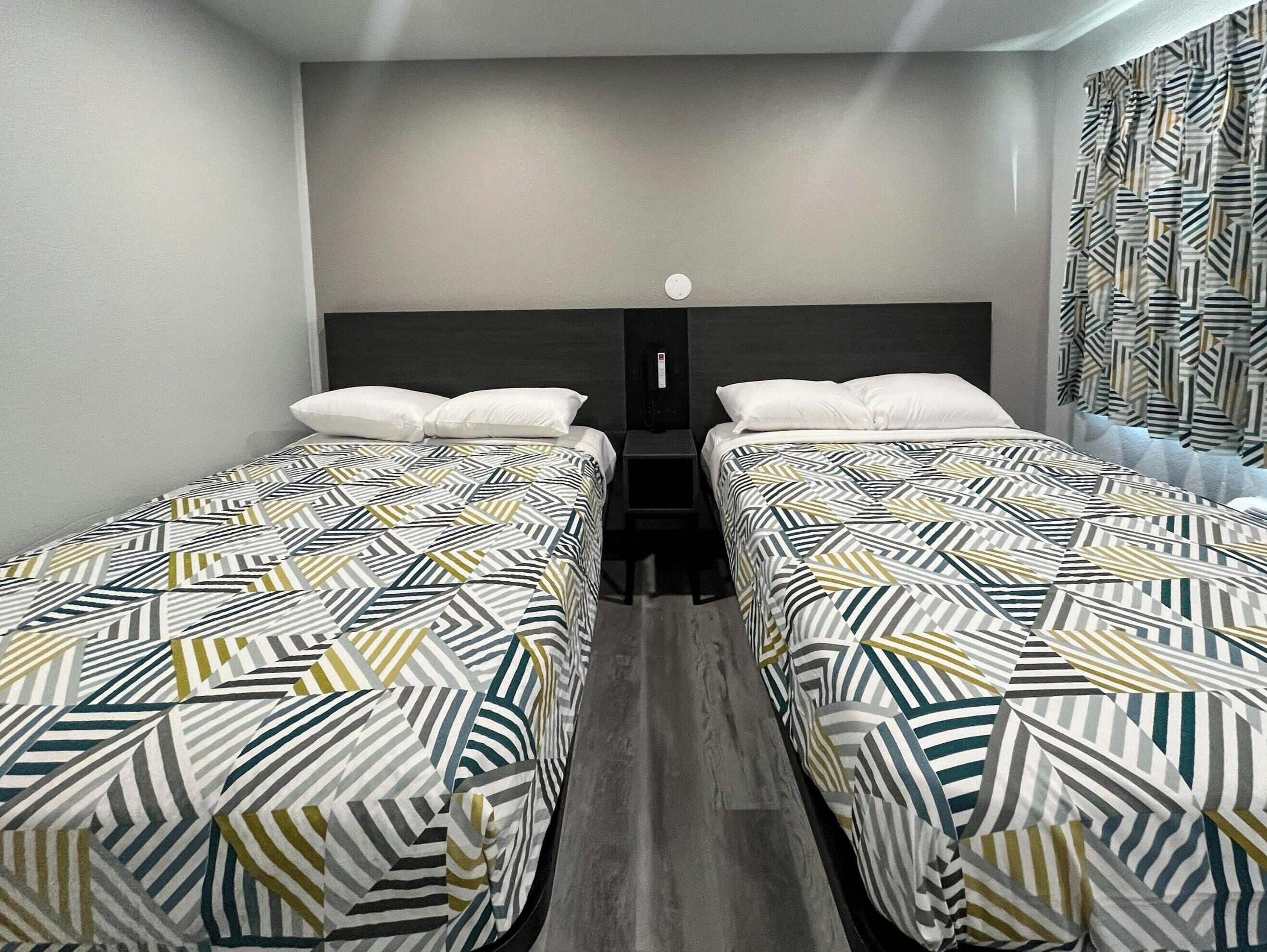 Habitación de hotel con dos camas individuales, una mesita de noche, una lámpara de pared y una cortina estampada.