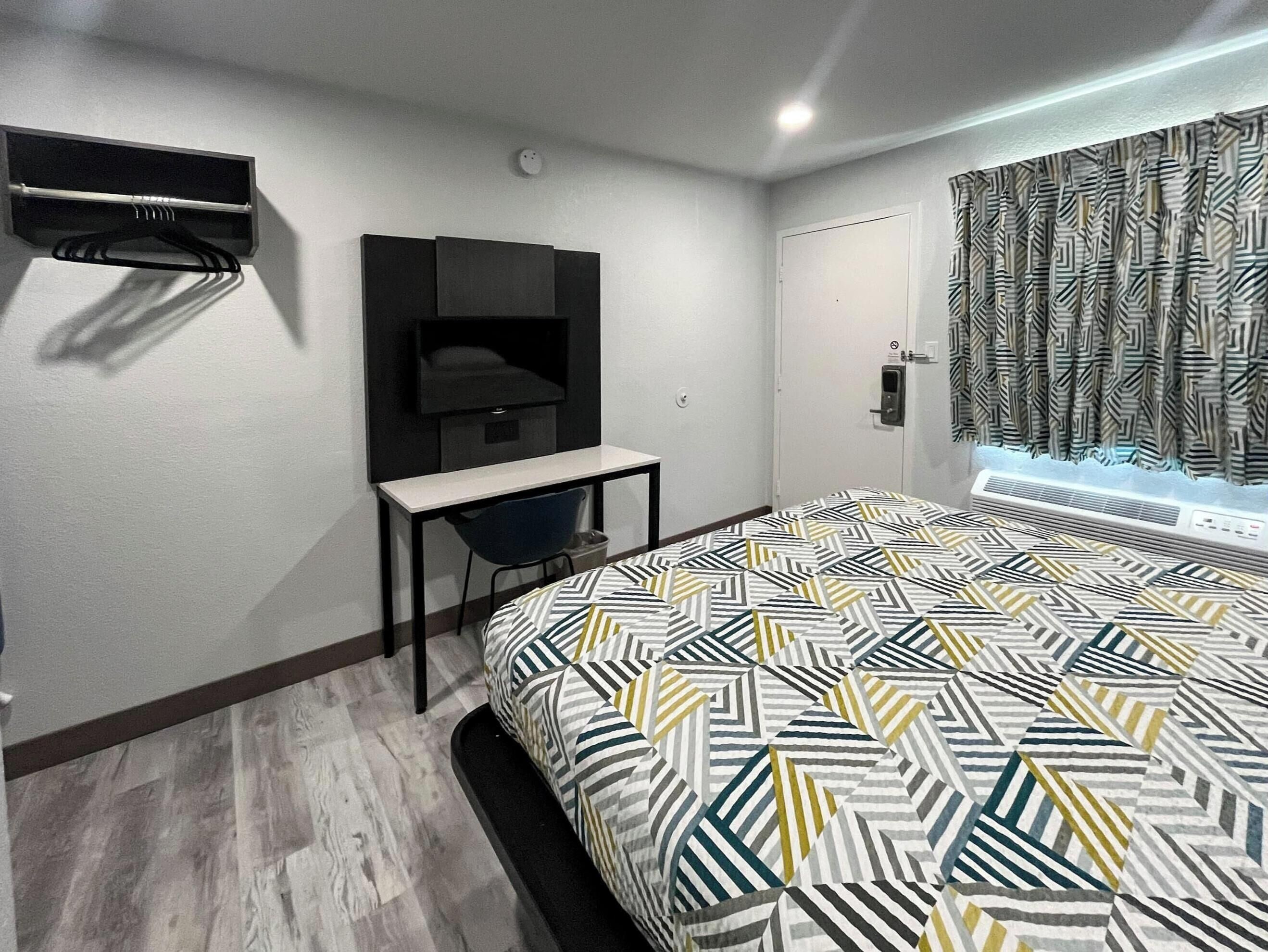 A modern hotel room with a bed, a desk, a TV, and a window with patterned curtains.